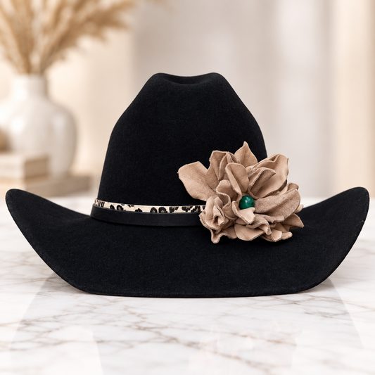 A black felt cowboy hat with a natural leather flower accent and a leopard-print hat band, displayed on a white marble surface with a blurred background.