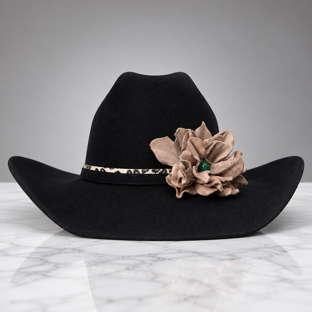 A black felt cowboy hat with a beige fabric flower accent and a leopard-print band, displayed on a white marble surface against a gray background.