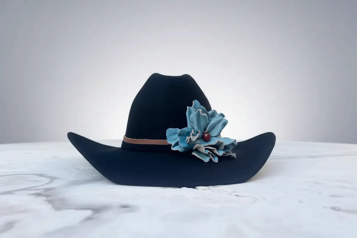 Black cowboy hat with a turquoise leather flower and a red agate stone on the band, displayed on a white marble surface against a gray background.