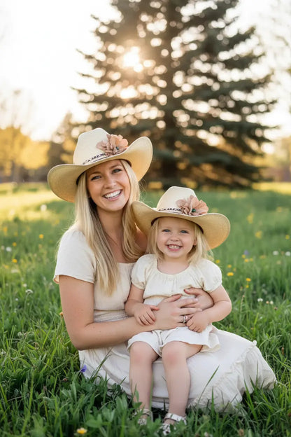 Woman and daughter wearing cowboy hats sitting in a grassy field with trees in the background. They are wearing Mommy and Me Cowgirl Hats with a beautiful matching leather flower leopard lace hat band. 