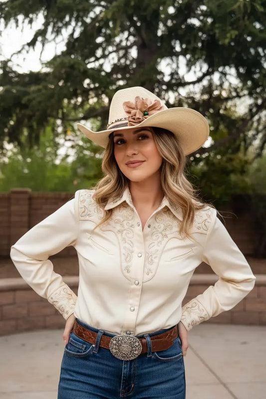 Woman wearing a cowgirl hat with a beautiful leather flower leopard lace hat band on it. She is also wearing a cream embroidered shirt, a belt buckle, and blue jeans.